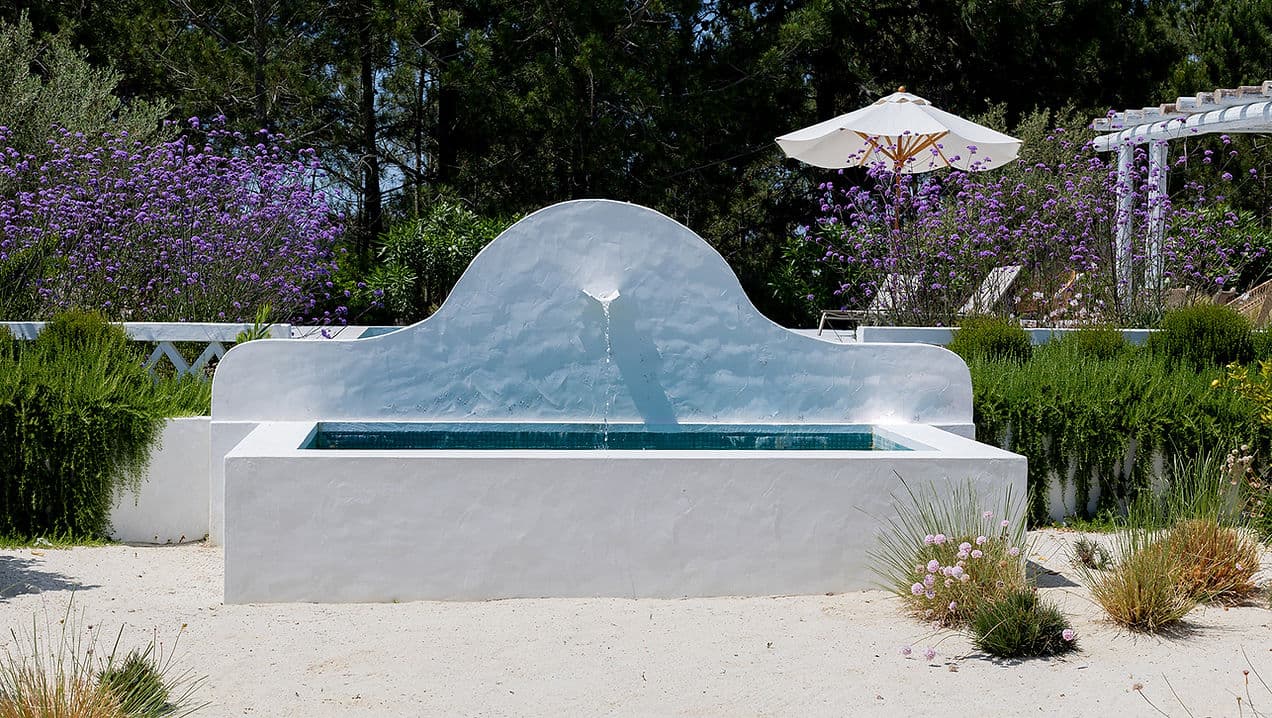 Outdoor white water fountain with greenery and purple flowers at Rafter Comporta Celeste in Comporta, Portugal