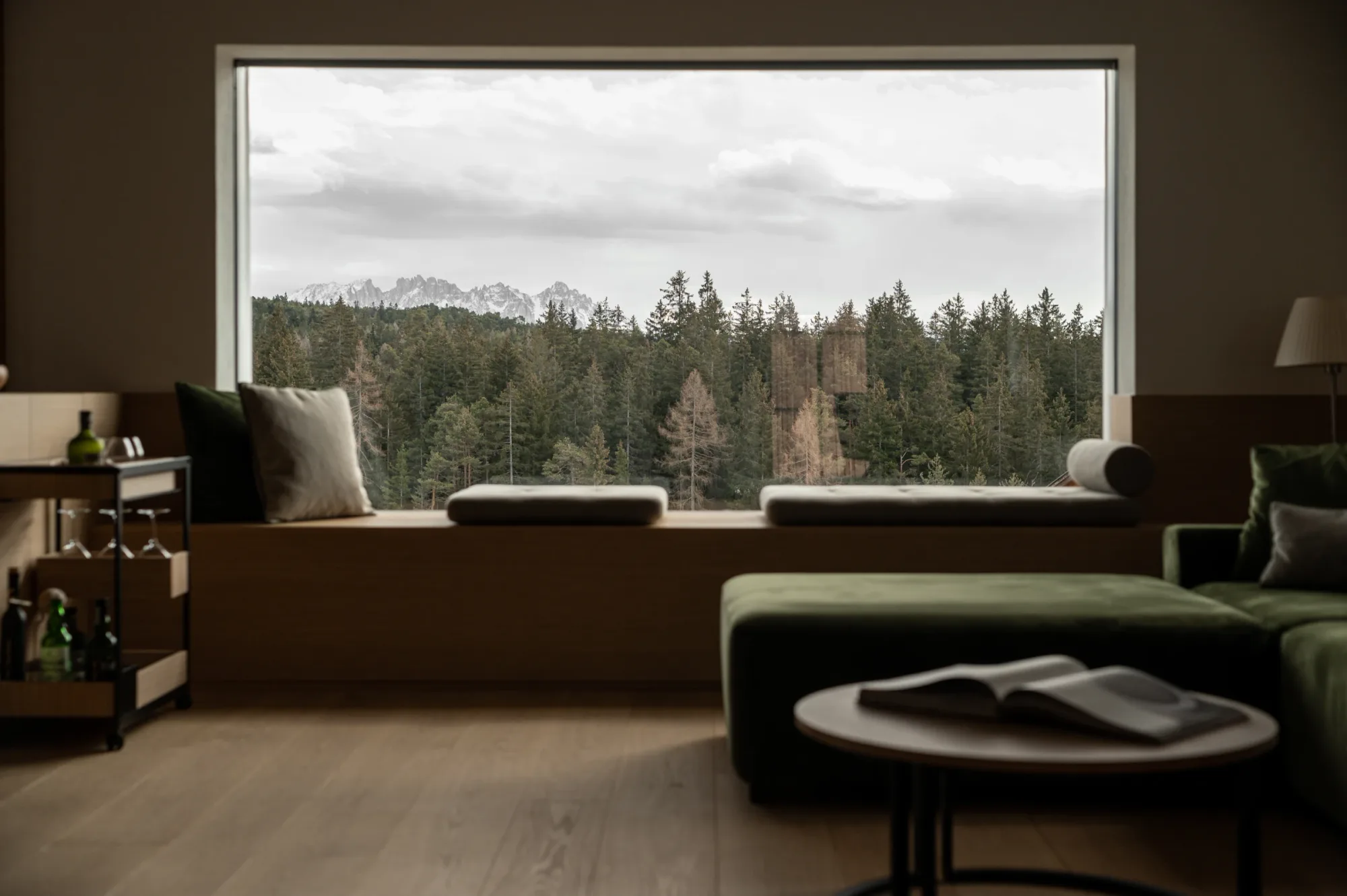 Living room with green sofa and floor-to-ceiling window framing forest and mountain view at Rafter Bolzano Oberinn in Italy