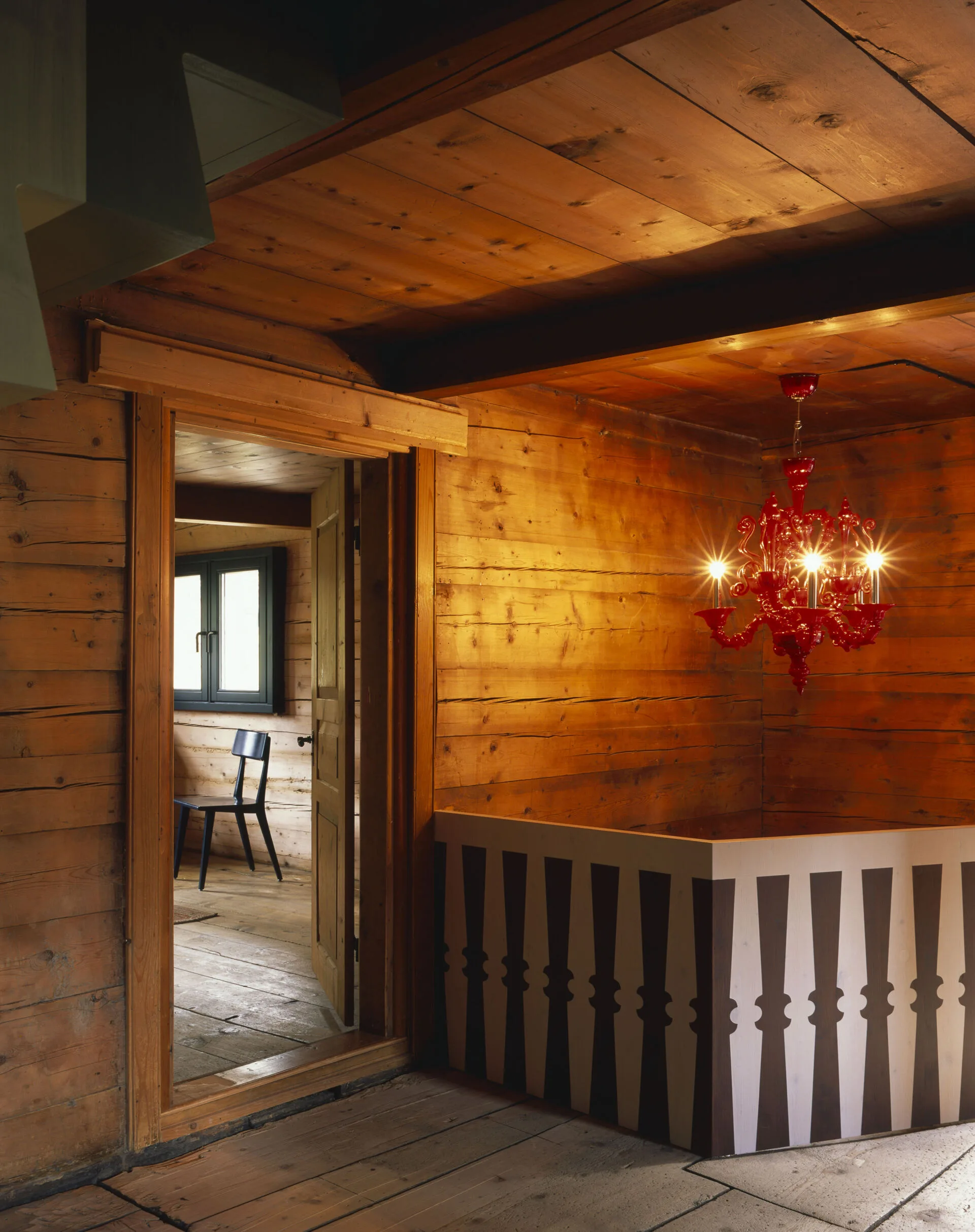 Rustic wooden interior with red chandelier and view into a room with black chair at Rafter Tschiertschen, Tschiertschen, Swit